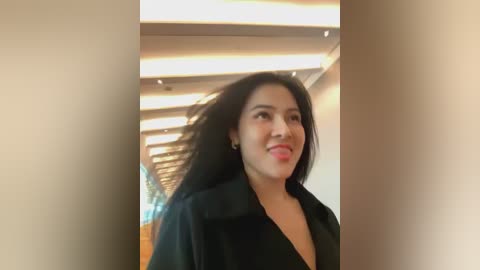 Media: Video of a smiling woman with long black hair and medium skin tone, wearing a black coat, in an indoor, modern setting with bright lights and white walls.