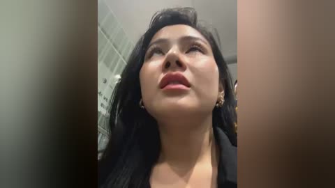 Media: A video of a young woman with medium skin tone, long black hair, and full lips, captured from a low angle. She wears a black outfit. Background features a blurry, tiled wall.