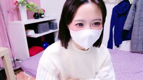 Media: Video of an Asian woman with straight, shoulder-length black hair, wearing a white face mask, white long-sleeve shirt, and light purple sweater. Background includes pink wall, white shelves, red and blue clothes, and a floral arrangement.