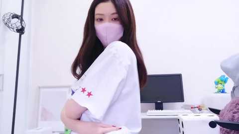 Media: Video of an Asian woman with long dark hair wearing a white mask and white shirt with red stars, standing in a white room with a desk and monitor.