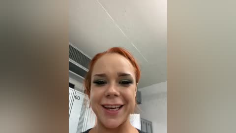 Media: Video of a young woman with fair skin and red hair, smiling, in a locker room with white lockers, an air conditioner, and a white ceiling.