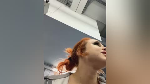 Media: A video of a pale-skinned woman with red hair in pigtails, wearing a gray hoodie, standing in a modern, industrial room with exposed ceiling beams and white walls.