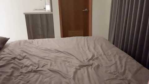 Media: A video of a neatly made bed with light gray sheets, a wooden door with a silver handle slightly ajar, and gray curtains in the background.