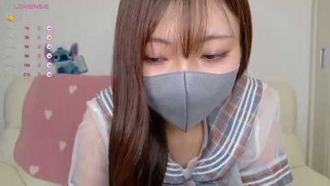 Media: A video of an Asian woman with straight brown hair, wearing a grey mask, a light blue and white striped shirt, and sitting on a pink heart-patterned cushion.