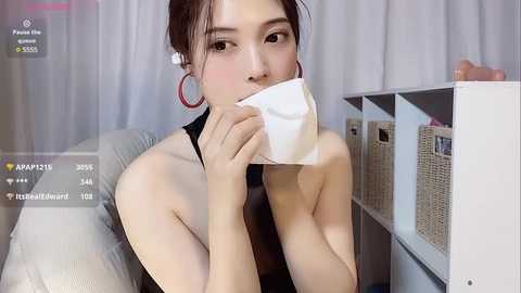 Media: Video of an East Asian woman with light skin, wearing a black sleeveless top, and large pink hoop earrings. She's blowing her nose into a tissue, with a shelf of wicker baskets in the background.