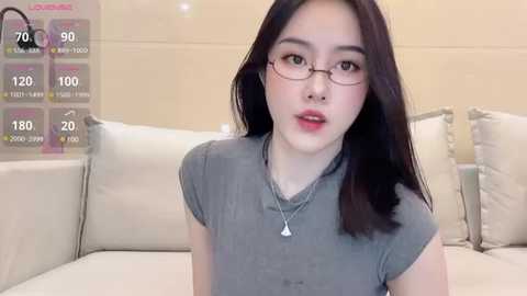 Media: A video of a young East Asian woman with fair skin, long black hair, and glasses, wearing a gray t-shirt, sitting on a white sofa in a modern living room.