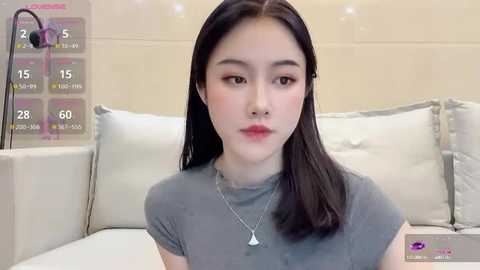 Media: A video of a young Asian woman with fair skin and long black hair, wearing a grey t-shirt and a silver necklace, sitting on a white couch.