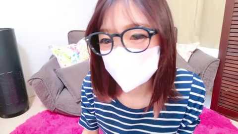 Media: Video of a young Asian woman with shoulder-length dark hair, wearing black-framed glasses and a white mask, seated on a pink rug in a room with beige walls, a gray couch, and a black floor lamp.