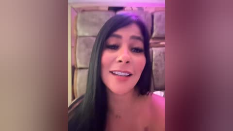 Media: Video of a Latina woman with long, dark hair and light skin, wearing minimal makeup, smiling. Background shows a rustic, stone wall with pink lighting.