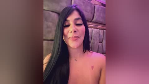 Media: Video of a topless Latina woman with long, straight black hair, closed eyes, and a subtle smile, standing against a rustic stone wall in a dimly lit room.