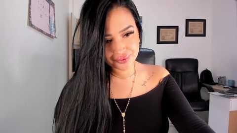 Media: Video of a smiling Latina woman with long black hair, wearing a black off-shoulder top, gold necklace, and a small tattoo on her right shoulder. She sits in an office with white walls, framed certificates, and a black leather chair.