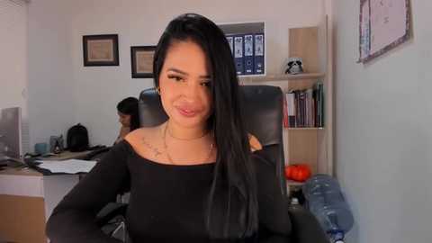 Media: Video of a smiling Latina woman with long black hair, wearing a black off-the-shoulder top, seated in an office with a framed certificate, a bookshelf, and a water dispenser.