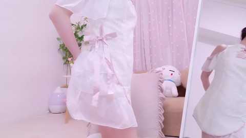 Media: A video of a slender woman in a white, short, sheer nurse dress with a pink ribbon, standing in a pastel-pink room with a plush toy and a vase on a white shelf.