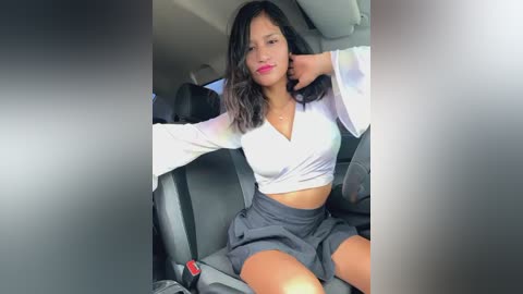 Media: Video of a young woman with medium skin tone, dark hair, wearing a white crop top and gray skirt, sitting in a car with black seats.