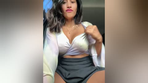 Media: Video of a Latina woman with medium-dark skin, wavy black hair, and full lips, wearing a white crop top and high-waisted black shorts, posing confidently indoors.