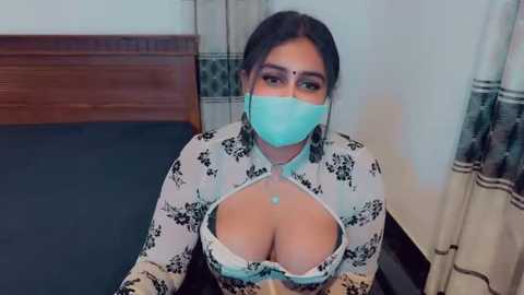 Media: Video of a woman with medium skin tone, dark hair, wearing a blue face mask, floral-patterned top revealing cleavage, sitting on a bed with a wooden headboard, beige curtains in the background.