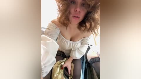 Media: Video of a young Caucasian woman with curly brown hair, wearing a white off-shoulder blouse, black stockings, and a gold handbag, sitting on a bed.