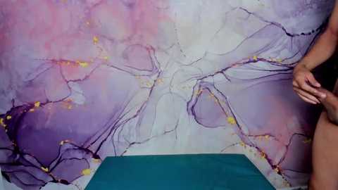 Media: Video of a person's lower torso and hand, standing in front of a vibrant, abstract mural with flowing purple and pink hues. The person wears black underwear, and the floor is covered in a green mat.