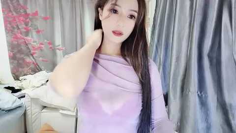 Media: Video of a fair-skinned, slender East Asian woman with long brown hair, wearing a sheer lavender top, posing against a backdrop of pink cherry blossoms and gray curtains.