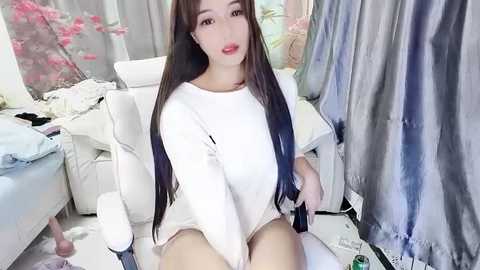 Media: A video of a young Asian woman with long, straight black hair and fair skin, wearing a white, loose-fitting top and no pants, sitting on a white chair in a cluttered room with pink cherry blossom decor.