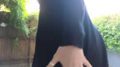 Media: Video of a person's lower back, wearing a black shirt, standing in a backyard with a wooden fence, lush greenery, and sunlight filtering through.