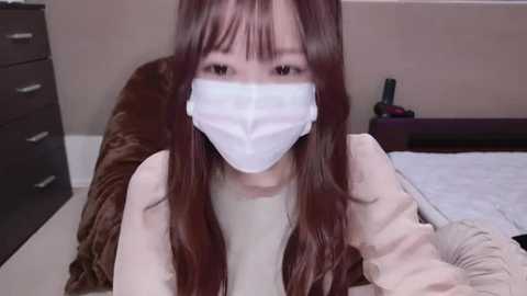 Media: Video of an Asian woman with long, straight brown hair and bangs, wearing a white face mask, beige sweater, and brown jacket. She is seated on a bed in a simple, beige room with dark wooden furniture.