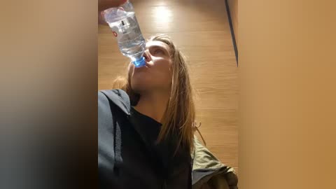 Media: A video captures a young woman with light brown hair, wearing a black blouse, drinking water from a clear plastic bottle on a wooden floor in a dimly lit room.