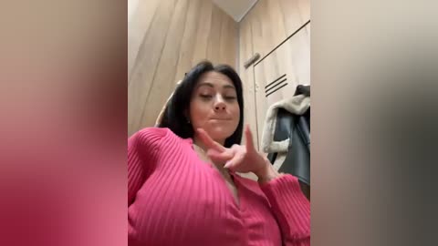 Media: Video of a Latina woman with long black hair, wearing a pink ribbed sweater, making a \"rock on\" gesture in a locker room with wooden lockers and a jacket hanging nearby.