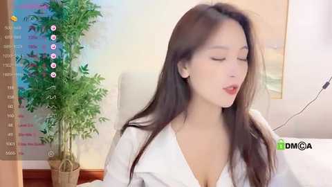 Media: A video of an East Asian woman with long black hair, wearing a white shirt, in a cozy room with a green plant and a calendar.