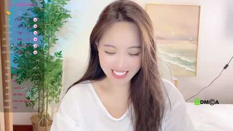 Media: Video of a smiling Asian woman with long brown hair, wearing a white top, in a brightly lit room with a potted plant and beach painting.