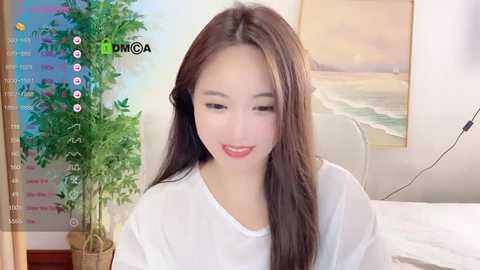 Media: A video of an Asian woman with long, straight, dark brown hair and fair skin, wearing a white shirt, smiling softly. Background features a potted plant, a framed beach painting, and a bed.