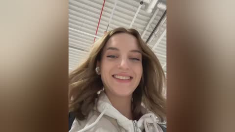 Media: Video of a smiling young woman with wavy light brown hair, wearing a light gray hoodie, standing in a modern, industrial-style room with white, ribbed ceiling panels and exposed pipes.