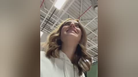 Media: A video of a young woman with wavy, light brown hair, wearing a white hoodie, smiling up towards the ceiling in a gymnasium with a corrugated metal roof and red ropes.