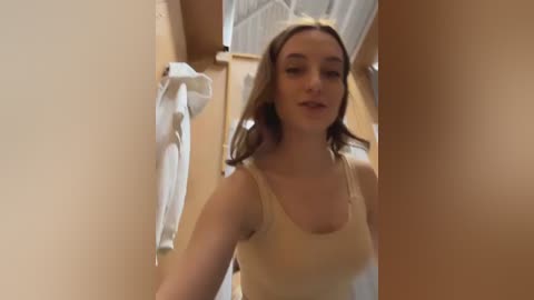 Media: Video of a young woman with light skin and straight, shoulder-length brown hair, wearing a beige tank top. She stands in a beige-walled hallway with a white towel hanging on a hook.