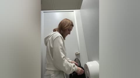 Media: Video of a young woman with shoulder-length brown hair in a white hoodie and gray sweatpants, standing in a narrow, white-tiled hallway, using a public restroom paper towel dispenser.