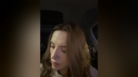 Media: Video of a young woman with light skin and long brown hair, wearing a white top, sitting in a dimly lit car, eyes closed, looking serene.