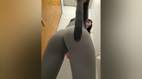 Media: Video of a woman in tight, grey leggings bending over, revealing her anus and erect penis. She has medium skin tone and long, dark hair. The background features a wooden door and white walls.