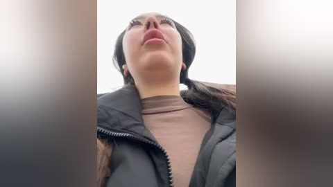 Media: Video of a woman with long, dark hair, wearing a black puffer jacket and a beige turtleneck, standing with her head tilted back, gazing upward.