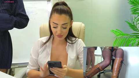 Media: Video of a light-skinned woman with dark hair, wearing a white blouse and brown leather boots, sitting in an office chair, texting, with a pinstriped man standing in the background.