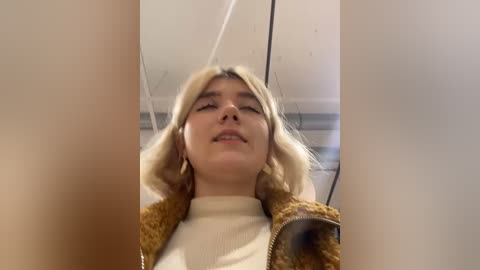 Media: Video of a young Caucasian woman with long, blonde hair, wearing a cream sweater and brown jacket, standing in a plain room with white ceiling and grey walls.