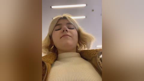 Media: A video of a young woman with shoulder-length blonde hair, wearing a beige ribbed sweater, and a brown fur-trimmed jacket, seen from a low angle, indoors with fluorescent lighting.