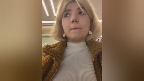 Media: A video of a young woman with short, blonde hair, wearing a ribbed white turtleneck under a brown, textured jacket, looking introspective with a neutral expression. The background is a blurred indoor setting with fluorescent lighting.