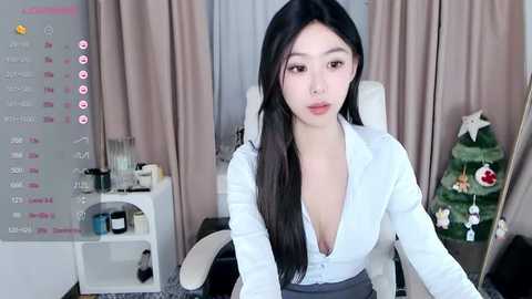 Media: A video of an Asian woman with long black hair, wearing a white blouse and gray skirt, sitting in an office chair in a room with beige curtains, a desk with a calendar, and a Christmas tree.