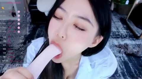 Media: Video of an Asian woman with long black hair, wearing a white shirt, giving a blowjob to a large, realistic, flesh-colored dildo. Background shows a cluttered room with a dark, patterned carpet and a potted plant.