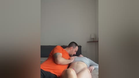 Media: Video of a muscular, tattooed man in an orange shirt kissing a pregnant woman's belly, both lying on a bed in a minimalistic room with beige walls.