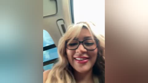 Media: Video of a smiling, light-skinned woman with wavy blonde hair, wearing black-rimmed glasses, inside a dimly lit train. The background shows a window with blue sky and a blurred train compartment.