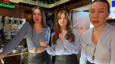 Media: Video of three young women in a bar, wearing light blue tops and black skirts. One woman ties her top, another holds a drink, and the third has a casual expression. Background shows a neon sign and bar decor.