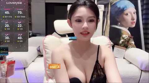 Media: A video of an East Asian woman with fair skin, dark hair, and wearing a black lace bra, sitting on a white chair in a modern living room with digital art and a painting in the background.