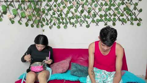 Media: Video of an Asian couple in a bright, modern room. Woman with straight black hair, black top, and pink shorts, plays with a pink doll. Man with short black hair, red tank top, and patterned shorts, sits nearby. Green vines and white flowers adorn the wall.