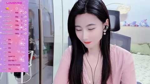 Media: Video of an East Asian woman with long black hair, fair skin, and red lipstick, wearing a pink top, sitting in a modern room with a colorful calendar, plush toys, and a TV.